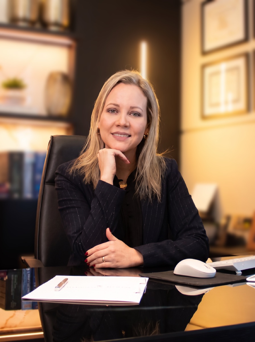 Carla Schons sentada à mesa do escritório, com a estante iluminada ao fundo
