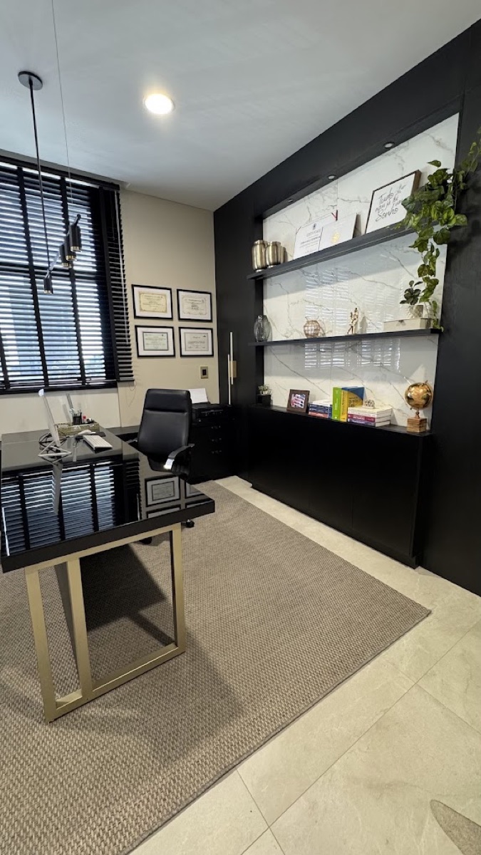 Sala do escritório: mesa preta com base dourada, estante iluminada e diplomas na parede